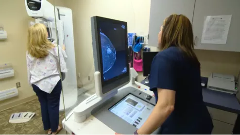 A mammography exam in progress with the patient on the breast imaging system and the technologist acquiring images. The image on the screen shows a suspect lesion.